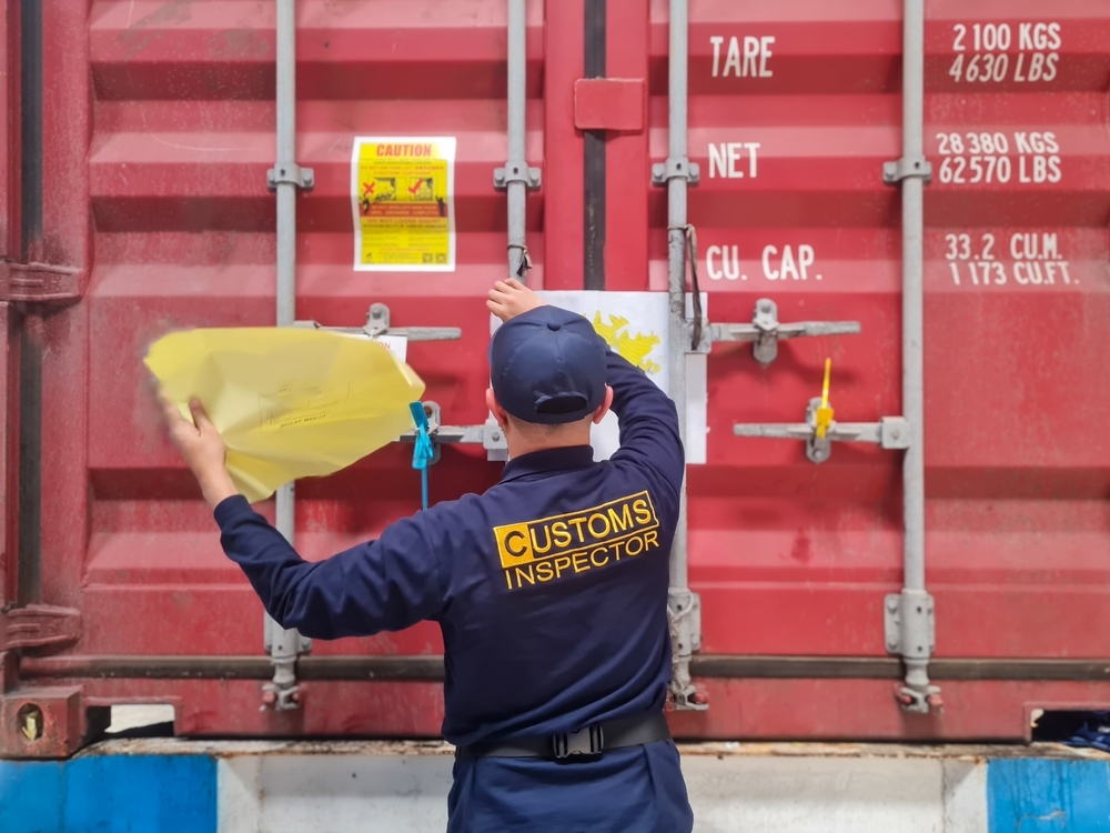 A customs inspector in uniform removes a yellow sticker from a red shipping container, which displays caution signs, weight labels, and secure metal locks—ensuring customs compliance essential for smooth global trade.