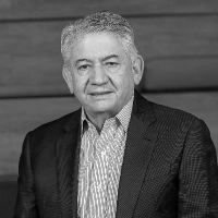 An older man in a suit jacket and striped shirt sits in front of a wooden background, looking at the camera.