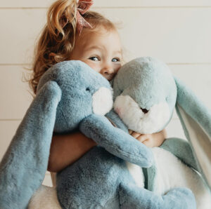 Smiling child hugs two large blue Bunnies by the Bay bunnies, with a bow in her hair.