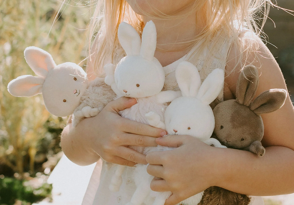 A child hugs four Bunnies by the Bay plush toys close to their chest outdoors.