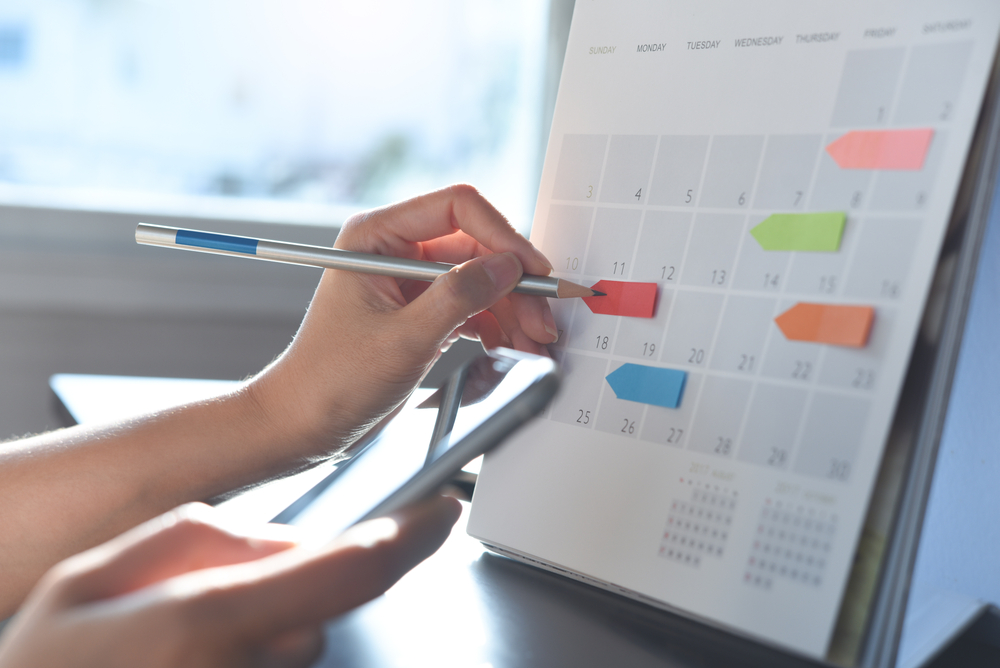 Hand pointing at a calendar with colored sticky notes, holding a pen and a smartphone.