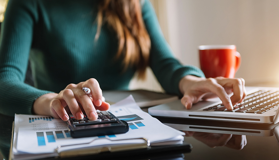 Person using a laptop and calculator to determine implied APR, surrounded by documents and a red coffee mug.