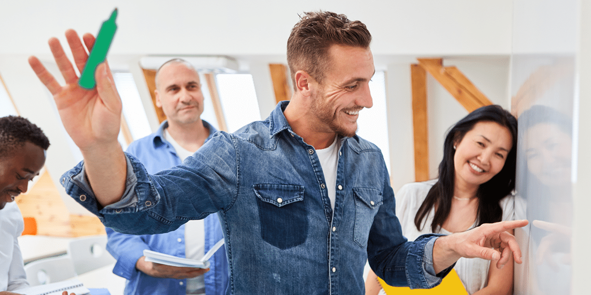 Man in denim shirt explains descuento dinámico on whiteboard as colleagues watch and smile in a bright meeting room.