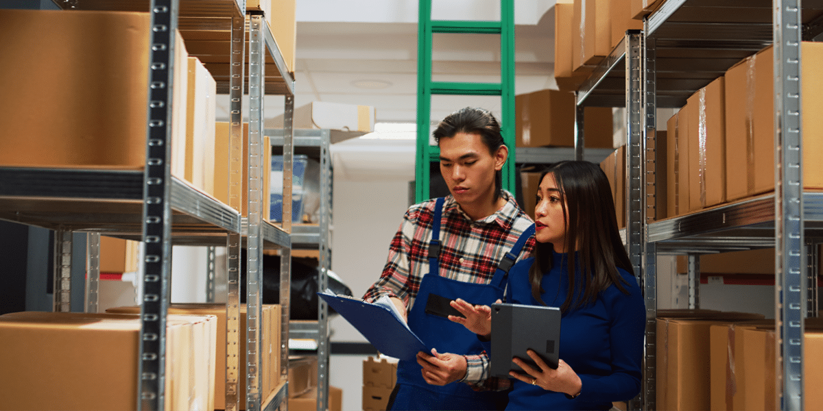 Two people checking inventory on shelves in a warehouse, holding a clipboard and a tablet.