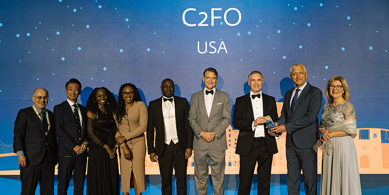 Nine people in formal attire stand on stage, smiling, at a C2FO USA event with a blue backdrop.
