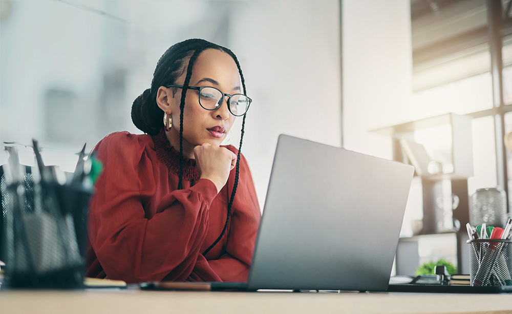 Woman with glasses and braids sits at a desk, thoughtfully browsing her summer reading book list on a laptop.
